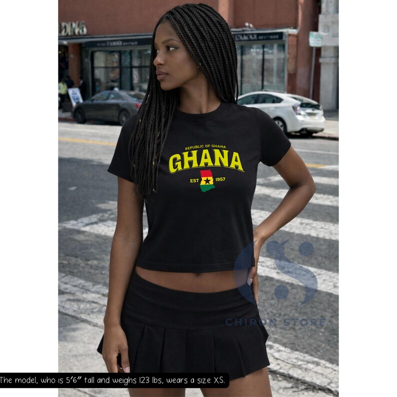 May include: A black short-sleeved crop top featuring the text Republic of Ghana GHANA EST 1957 in yellow and white, with a graphic of the Ghana flag in the center. The shirt is paired with a black pleated mini skirt.