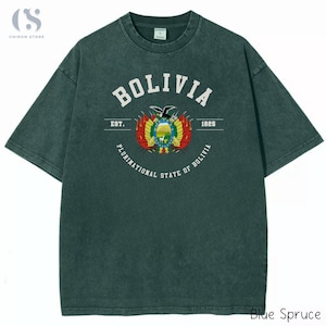 Puede incluir: Camiseta azul abeto con la palabra "BOLIVIA" en letras blancas en bloque sobre el escudo de armas boliviano. El texto "EST. 1825" está debajo de la palabra "BOLIVIA". Las palabras "PLURINATIONAL STATE OF BOLIVIA" están arqueadas debajo.