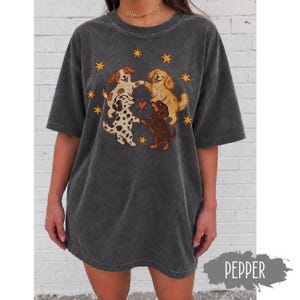以下が含まれることがあります： チャコールグレーの半袖Tシャツ。前足をつないで円を描く4匹の犬のグラフィックプリントが施されています。犬は茶色、白、斑点模様で、中央に小さな赤いハートがあります。犬の周りには黄色い四芒星が散りばめられています。