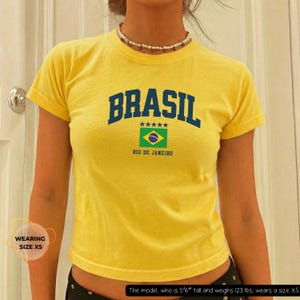 May include: Yellow t-shirt with the word "BRASIL" in navy blue block letters. Below the word is a graphic of the Brazilian flag and the text "RIO DE JANEIRO". The shirt is size XS.