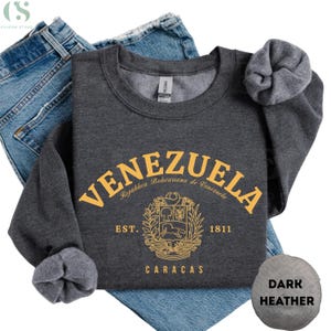May include: Dark heather gray sweatshirt with the word "VENEZUELA" in gold lettering. Below the word is a gold emblem and the words "EST. 1811 CARACAS". The sweatshirt is folded on top of blue jeans.