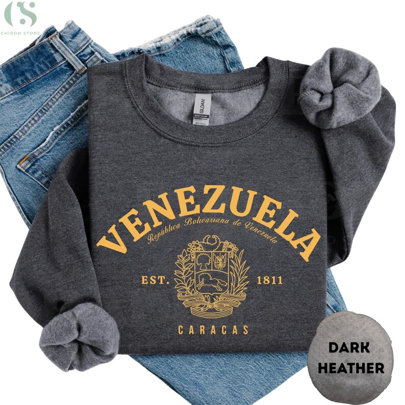 May include: Dark heather gray sweatshirt with the word "VENEZUELA" in gold lettering. Below the word is a gold emblem and the words "EST. 1811 CARACAS". The sweatshirt is folded on top of blue jeans.