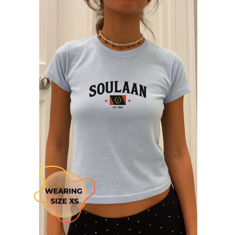 May include: Light blue t-shirt with the word "SOULAAN" in black, a logo, and "EST. 1865". The shirt is short-sleeved. The person is wearing a necklace. The text "WEARING SIZE XS" is in a yellow circle.