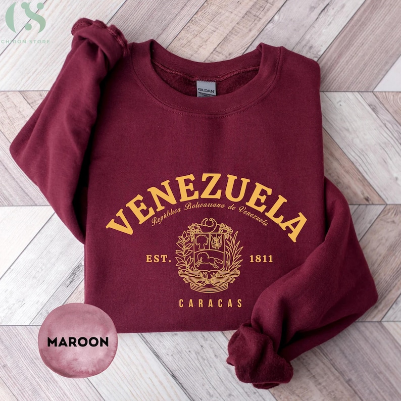 May include: Maroon sweatshirt with the word "VENEZUELA" in gold arched lettering. Below the text is a gold emblem with the words "Rep&uacute;blica Bolivariana de Venezuela", "EST. 1811", and "CARACAS". The sweatshirt is folded on a light wood surface.