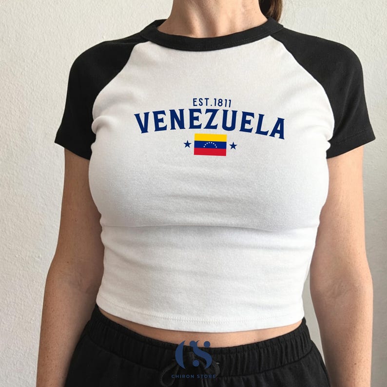 May include: White crop top with black raglan sleeves featuring the word "VENEZUELA" in navy blue, the Venezuelan flag, and the text "EST.1811". The shirt is a casual style.