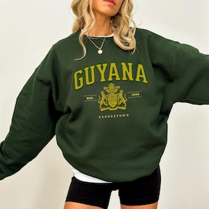 Peut inclure: Sweat-shirt vert foncé avec le mot "GUYANA" en lettres dorées. En dessous, un blason avec le texte "EST. 1966" et "GEORGETOWN". Le sweat-shirt est de style col rond et a des manches longues.