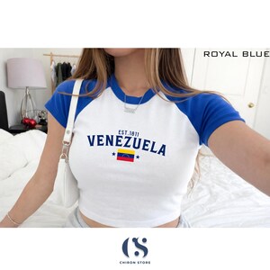 May include: White and royal blue raglan crop top with the word "VENEZUELA" printed on the front, along with the Venezuelan flag and the text "EST. 1811". The shirt has short sleeves and a round neckline.