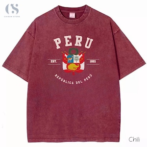 May include: A maroon t-shirt with a distressed finish. The front features the word "PERU" above a crest with a deer, tree, and other symbols. Below the crest is "REPUBLICA DEL PERU" and "EST. 1821".
