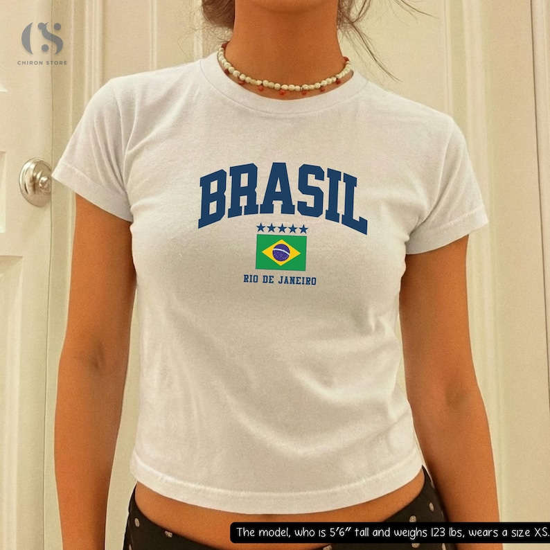 May include: White t-shirt with the word "BRASIL" in blue block letters, a Brazilian flag graphic, and "RIO DE JANEIRO" printed below. The shirt has short sleeves and a crew neck.