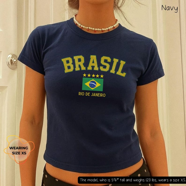 May include: Navy blue crop top with the word "BRASIL" in yellow block letters, a Brazilian flag graphic, and the text "RIO DE JANEIRO". The shirt has short sleeves and a crew neck. The model is wearing size XS.