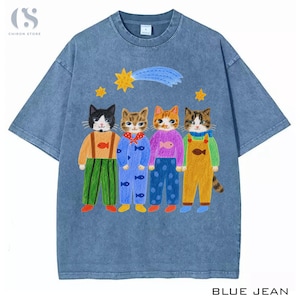May include: A blue acid-wash t-shirt featuring a graphic of four anthropomorphic cats wearing colorful clothing. Above the cats, there is a yellow shooting star and two smaller stars. The cats are depicted in various outfits including overalls and patterned shirts with fish motifs.