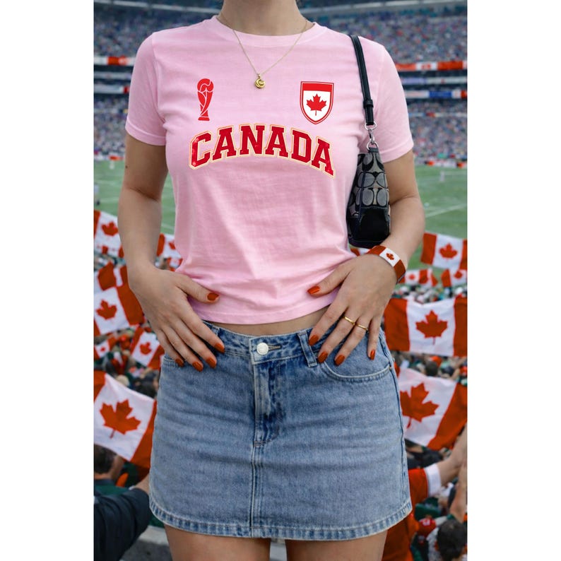 Peut inclure: Un t-shirt rose clair avec le mot "CANADA" en rouge, ainsi qu'un embl&egrave;me de feuille d'&eacute;rable rouge. La chemise est associ&eacute;e &agrave; une jupe en jean. Un sac &agrave; main noir et un bracelet avec un motif de feuille d'&eacute;rable sont &eacute;galement visibles.