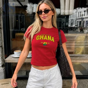 May include: A red short-sleeved t-shirt featuring the text Republic of Ghana and Ghana in yellow letters, with a small graphic of the Ghana flag and the text EST 1957. The shirt is paired with white pants and a dark brown woven shoulder bag.