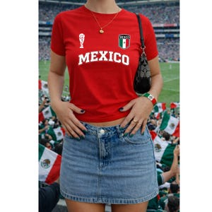 Op de afbeelding: Een rood T-shirt met korte mouwen met het woord MEXICO in witte letters, een klein wit trofee-icoon en een Mexico-vlagembleem op de borst. Het shirt wordt gecombineerd met een blauwe spijkerrok met een zichtbare zilveren knoop.