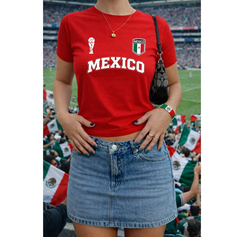 Op de afbeelding: Een rood T-shirt met korte mouwen met het woord MEXICO in witte letters, een klein wit trofee-icoon en een Mexico-vlagembleem op de borst. Het shirt wordt gecombineerd met een blauwe spijkerrok met een zichtbare zilveren knoop.