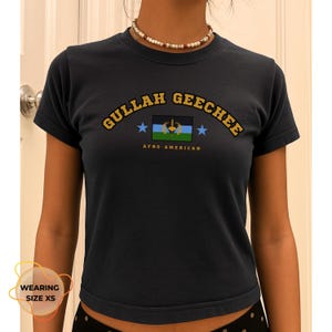 May include: Dark navy blue t-shirt featuring the text "GULLAH GEECHEE" in yellow arched lettering. Below the text is a graphic of a flag with blue, green, and black stripes, and two blue stars. The text "AFRO-AMERICAN" is below the flag.