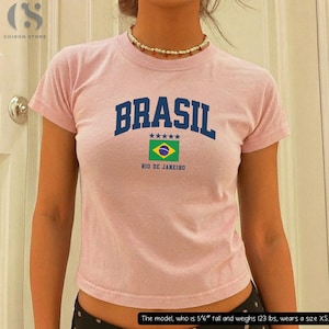 May include: A light pink t-shirt with the word "BRASIL" in navy blue block letters, a Brazilian flag graphic, and "RIO DE JANEIRO" underneath. The shirt is short-sleeved and has a crew neck.