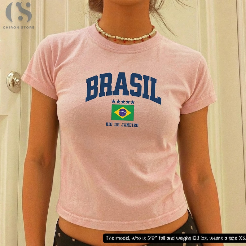 May include: A light pink t-shirt with the word "BRASIL" in navy blue block letters, a Brazilian flag graphic, and "RIO DE JANEIRO" underneath. The shirt is short-sleeved and has a crew neck.