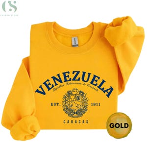 May include: A gold-colored sweatshirt with the word "VENEZUELA" in navy blue arched across the front. Below the word is a crest and the words "EST. 1811 CARACAS". The sweatshirt has a round neck and long sleeves.