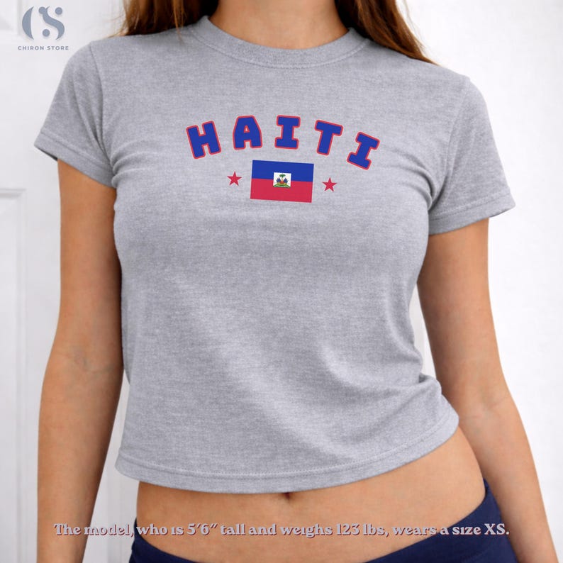May include: A gray crop top with the word "HAITI" in blue letters and a Haitian flag graphic. The shirt has a crew neck and short sleeves. The model is wearing a size XS.