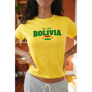 Puede incluir: Camiseta amarilla con la palabra "BOLIVIA" en letras verdes, el texto "Est. 1821" arriba y la bandera boliviana debajo. La camiseta es de manga corta y tiene cuello redondo.