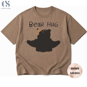 May include: A brown t-shirt featuring the words "Bear Hug" and a black bear graphic with open arms. The shirt is a neutral brown colour, and the bear has a tan hat. The t-shirt is a casual style.