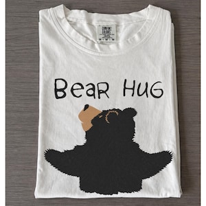 Può includere: T-shirt bianca con la scritta nera "Bear Hug" e un'illustrazione di un orso nero con muso marrone e braccia aperte. La maglietta è piegata ordinatamente su una superficie di legno.