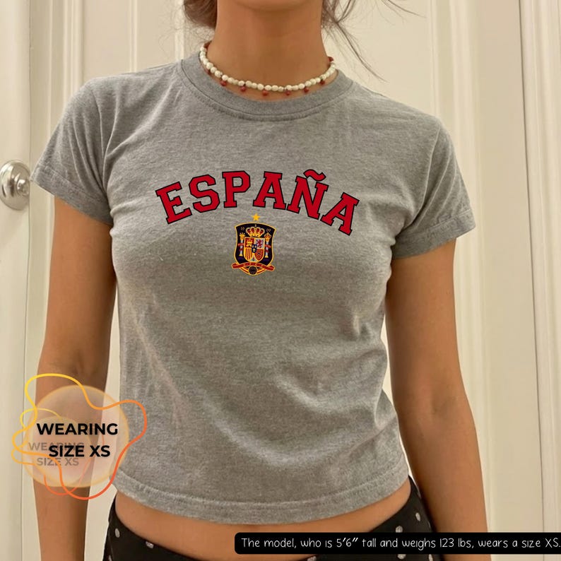 May include: A gray, short-sleeved t-shirt with the word "ESPA&Ntilde;A" in red arched lettering across the chest. Below the text is a crest with a crown and shield. The model is wearing size XS.
