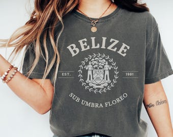 May include: Dark gray t-shirt with the word "BELIZE" in a vintage font above a circular emblem. The emblem includes the text "EST. 1981" and the Latin phrase "SUB UMBRA FLOREO". The shirt has short sleeves and a crew neck.