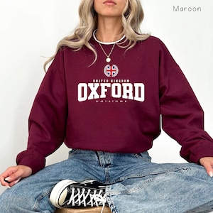 以下が含まれることがあります： 白い文字で「OXFORD」と書かれたマルーン色のスウェットシャツ。上部には「UNITED KINGDOM」の文字と、ユニオンジャックの紋章があります。スウェットシャツは無地のマルーン色です。