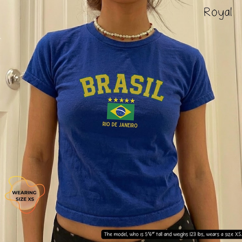 May include: Royal blue t-shirt with the word "BRASIL" in yellow block letters. Below the word are five yellow stars and a Brazilian flag graphic with "RIO DE JANEIRO" underneath. The shirt is a cropped style.