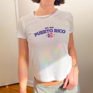 以下が含まれることがあります： 前面に「EST. 1508 PUERTO RICO」の文字と、青と赤のプエルトリコの国旗のグラフィックがプリントされた白いTシャツです。