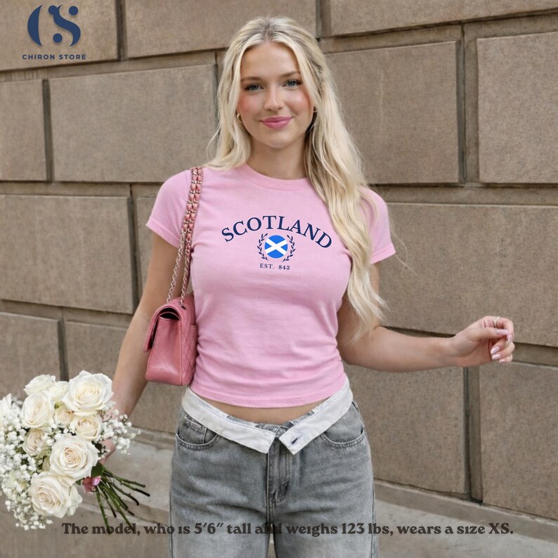 Peut inclure: T-shirt rose avec le mot "SCOTLAND" en arc de cercle, un embl&egrave;me bleu et blanc et le texte "EST. 848". Le mod&egrave;le porte un jean gris. Le mod&egrave;le mesure 167 cm et p&egrave;se 55,7 kg.