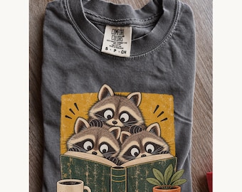 May include: A charcoal gray t-shirt with a Comfort Colors label at the neck. The graphic features three raccoons reading a large green book on a yellow background, accompanied by a small potted plant and a coffee mug in the foreground.