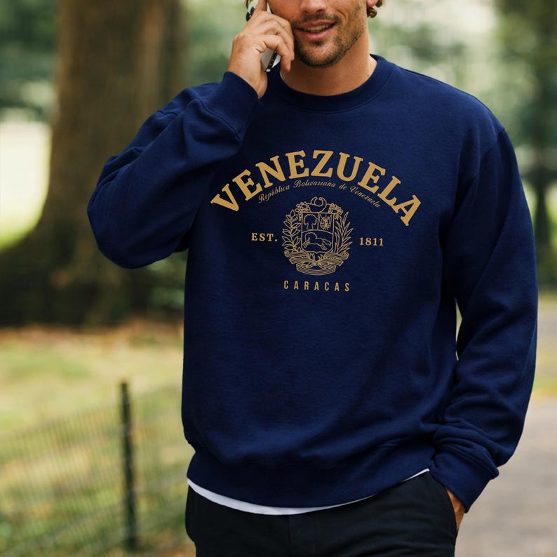 May include: Navy blue sweatshirt with gold lettering and a crest design. The word "VENEZUELA" arches across the top, with "Rep&uacute;blica Bolivariana de Venezuela" above the crest. Below the crest are the words "EST. 1811 CARACAS".