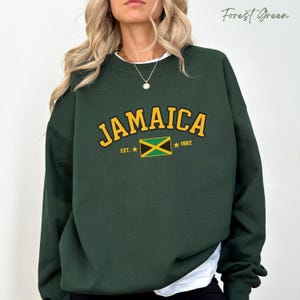 Op de afbeelding: Bostgroen sweatshirt met "JAMAICA" in gouden letters. Het ontwerp bevat de Jamaicaanse vlag, het jaar "1962" en de tekst "EST.". De sweatshirt heeft een ronde hals.