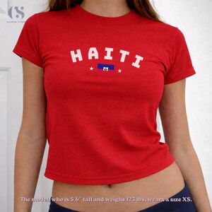 May include: Red crop top with the word "HAITI" in white block letters across the front. The shirt features a small Haitian flag graphic and two white stars. The shirt is a crew neck and has short sleeves.
