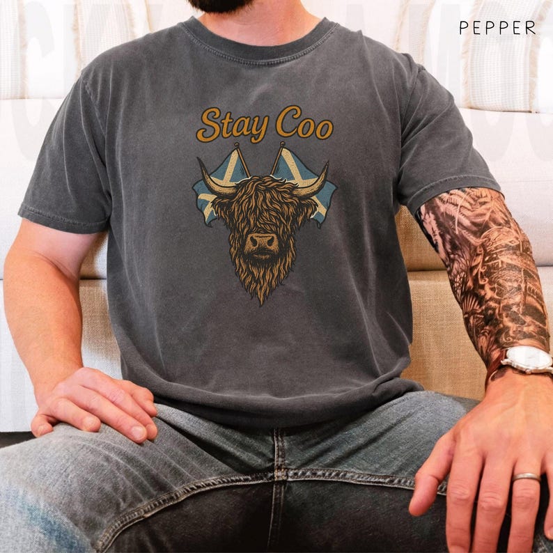 K&ouml;nnte beinhalten: Dunkelgraues T-Shirt mit einer Grafik einer Highland-Kuh, schottischen Flaggen und dem Text "Stay Coo" in Gold. Das Design zeigt eine detaillierte Illustration des Gesichts und der H&ouml;rner der Kuh. Ein l&auml;ssiges Kleidungsst&uuml;ck.