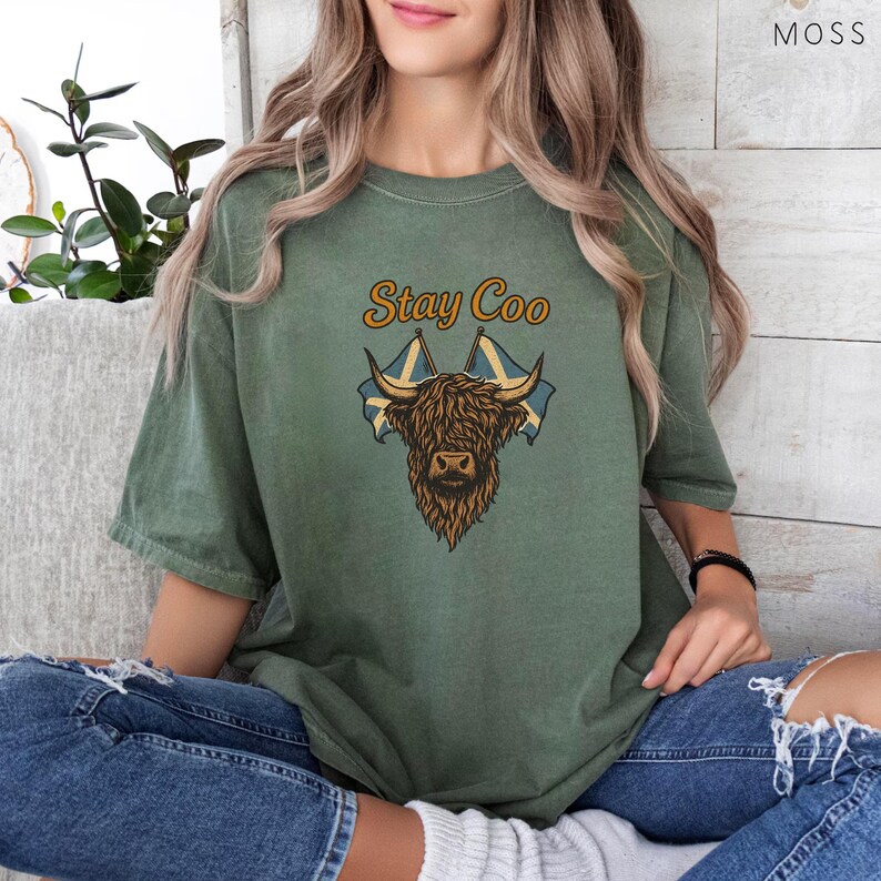 K&ouml;nnte beinhalten: Olivgr&uuml;nes T-Shirt mit dem Text "Stay Coo" &uuml;ber einer braunen Highland-Kuh-Grafik und zwei schottischen Flaggen. Das Shirt hat eine lockere Passform und kurze &Auml;rmel.