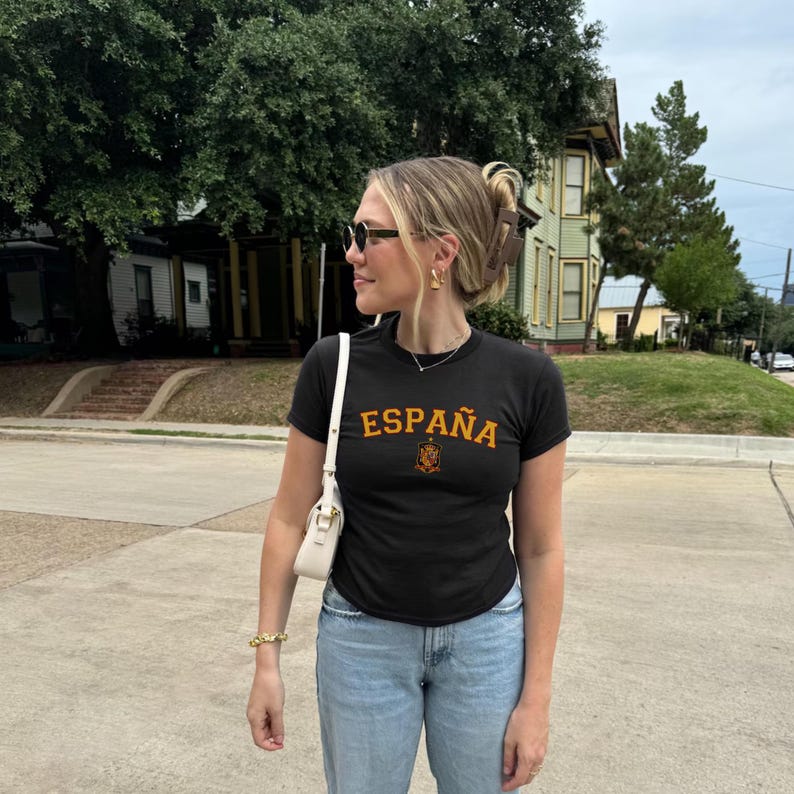 May include: Black t-shirt with the word "ESPA&Ntilde;A" in orange block letters and a small emblem. The shirt is paired with light blue jeans and a white purse. The person is wearing sunglasses and gold jewelry.