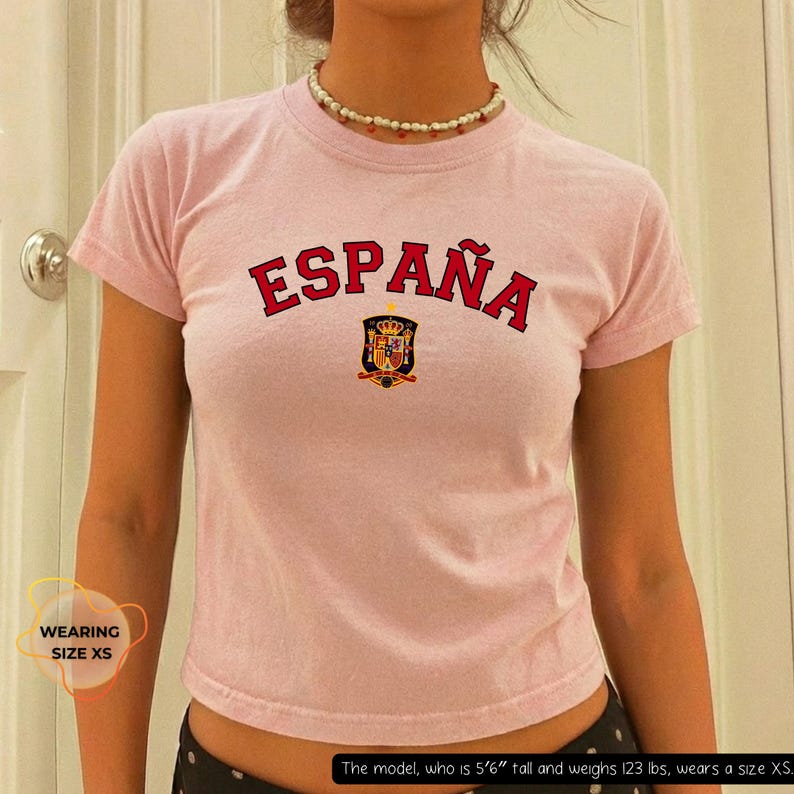 May include: A light pink t-shirt with the word "ESPA&Ntilde;A" in red block letters across the chest. Below the text is a crest with a crown and shield. The shirt is short-sleeved and has a crew neck.