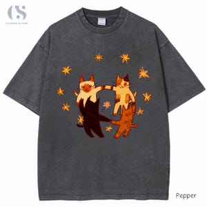 Op de afbeelding: Een antracietgrijs t-shirt met korte mouwen en een grafische print van vier katten in verschillende kleuren die elkaar in een cirkel bij de pootjes vasthouden, omringd door kleine oranje sterren. De stof heeft een verwassen look en een ronde hals.