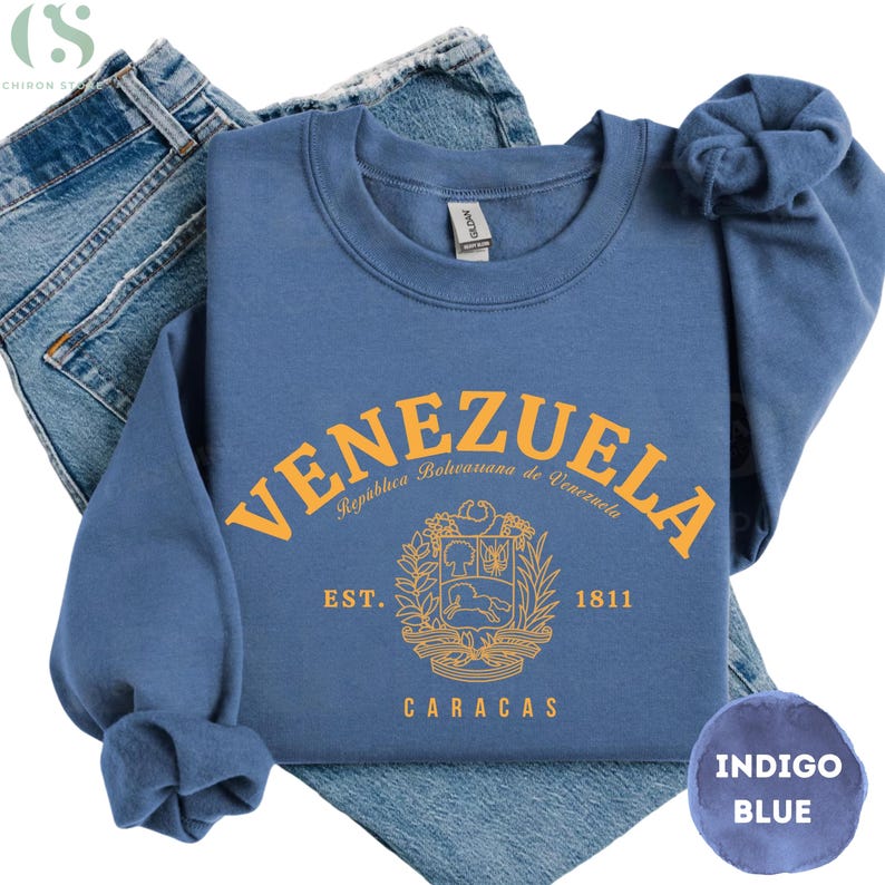 May include: Indigo blue sweatshirt with the word "VENEZUELA" in gold lettering. The sweatshirt features a crest design and the text "CARACAS" and "1811". The sweatshirt is folded on top of a pair of blue jeans.