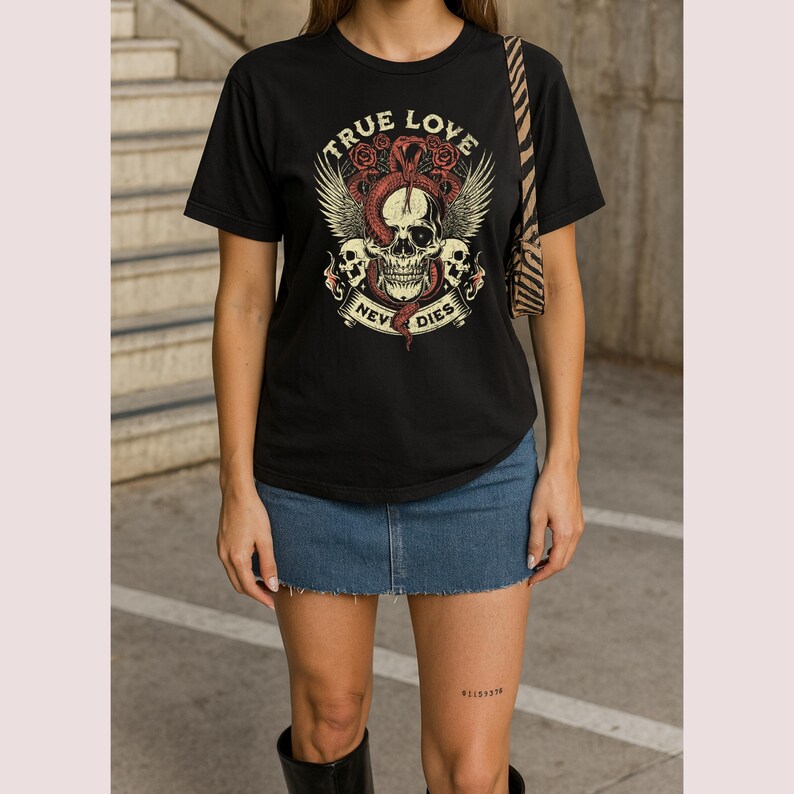 Pode incluir: Camiseta preta com um gr&aacute;fico de caveira, asas, rosas e uma cobra. O texto "TRUE LOVE" est&aacute; acima da caveira, com "NEVER DIES" em uma faixa abaixo. A camiseta &eacute; combinada com uma saia jeans e botas pretas.