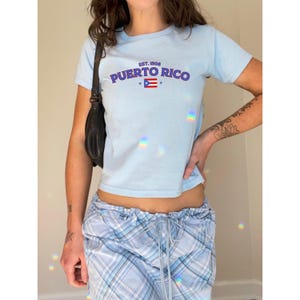 以下が含まれることがあります： 薄い青色のクロップドTシャツに、紫色の「EST. 1908 PUERTO RICO」の文字と、プエルトリコの国旗の小さな画像がプリントされています。