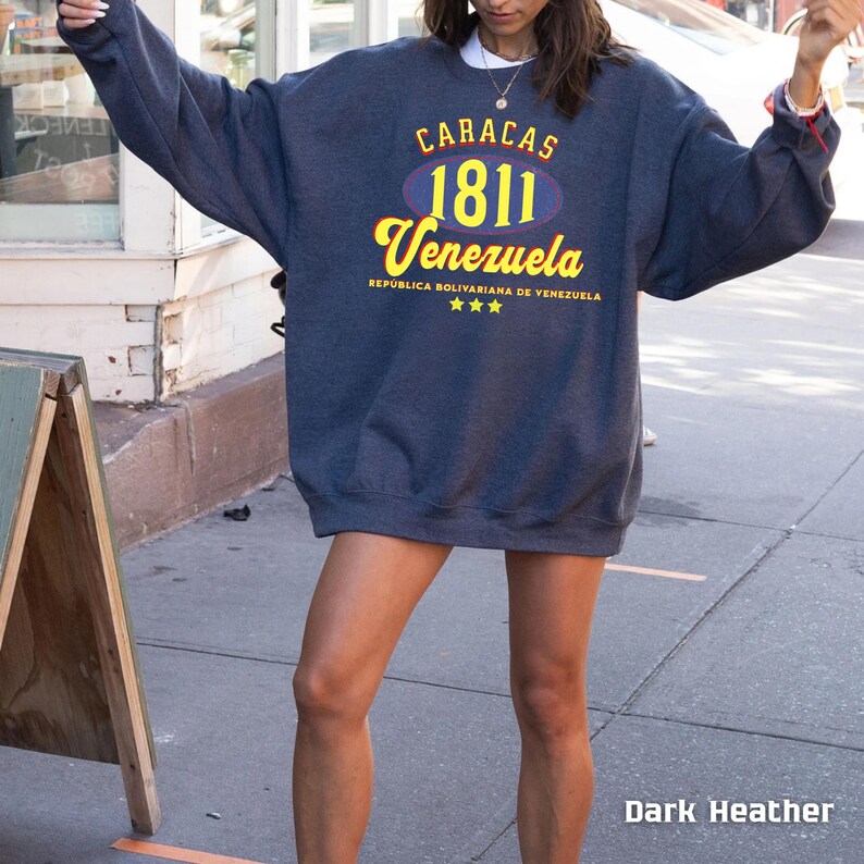 May include: Dark heather gray sweatshirt with the text "CARACAS 1811 Venezuela" in yellow and blue. The text "REPUBLICA BOLIVARIANA DE VENEZUELA" is below the word "Venezuela". The sweatshirt is oversized.