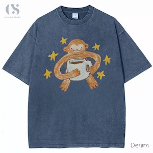 以下が含まれることがあります： コーヒーカップを持った猿の漫画が描かれた、青いデニムのTシャツ。黄色い星が周りに描かれています。Tシャツはクルーネックで半袖です。「Denim」の文字が右下にプリントされています。