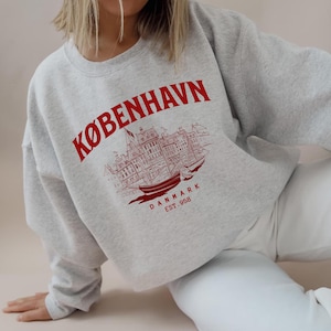 Könnte beinhalten: Ein hellgraues Sweatshirt mit dem Wort "KØBENHAVN" in roter Schrift, das sich oben wölbt. Darunter befindet sich eine rote Illustration einer Stadtlandschaft und eines Schiffs. Der Text "DANMARK EST. 958" ist darunter gedruckt.