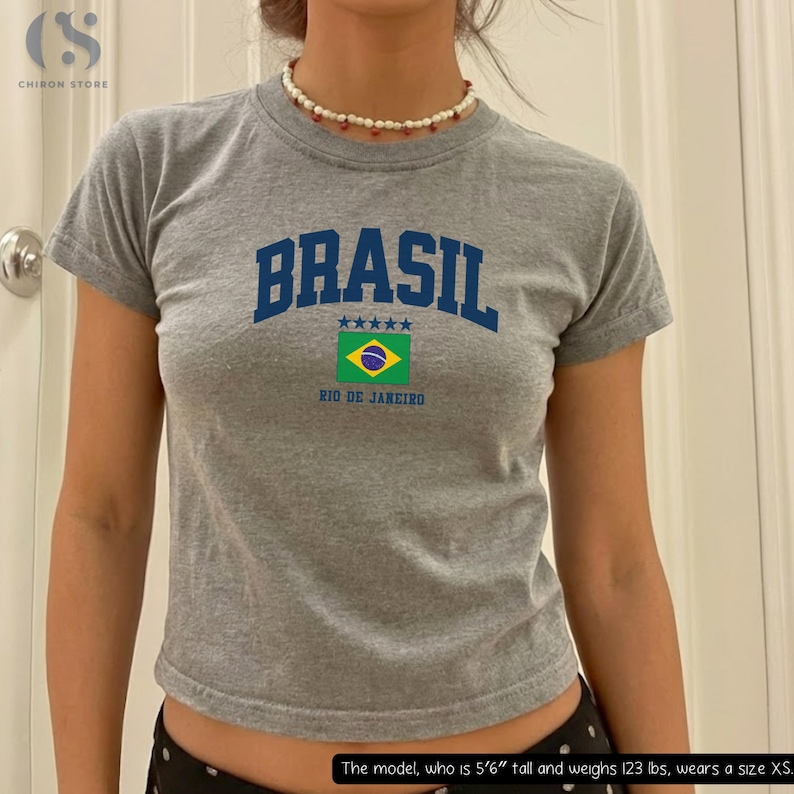 May include: A gray t-shirt with the word "BRASIL" in blue block letters, a Brazilian flag graphic, and the text "RIO DE JANEIRO." The shirt has short sleeves.