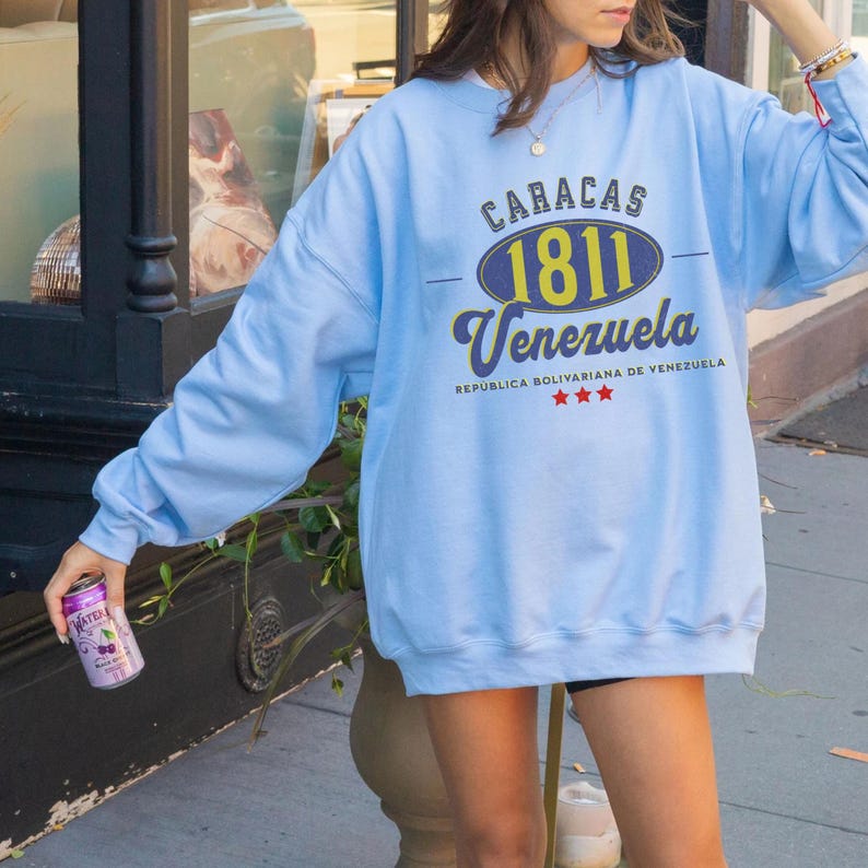 May include: A light blue sweatshirt with the text "CARACAS 1811 Venezuela" in navy blue, yellow, and white. The sweatshirt also has the text "REPUBLICA BOLIVARIANA DE VENEZUELA" and three red stars. The person is holding a can of beverage.